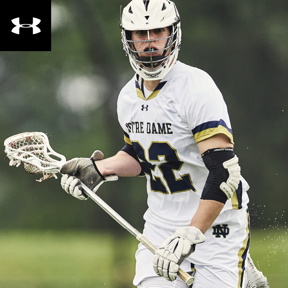 A player wearing a under armour lacrosse uniform is on the field equipped with a helmet, elbow pads and gloves is cradling a lacrosse stick with a ball in the netting.