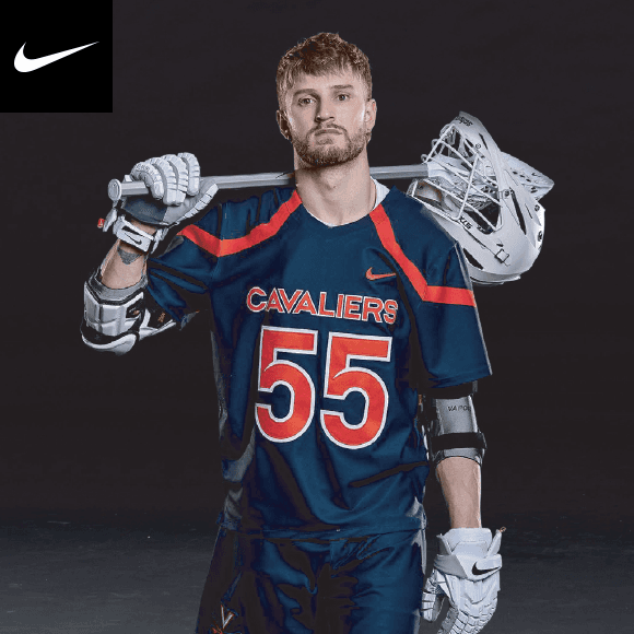 Player wearing elbow pads and gloves is standing wearing a Nike lacrosse uniform with lacrosse stick and helmet over his shoulder